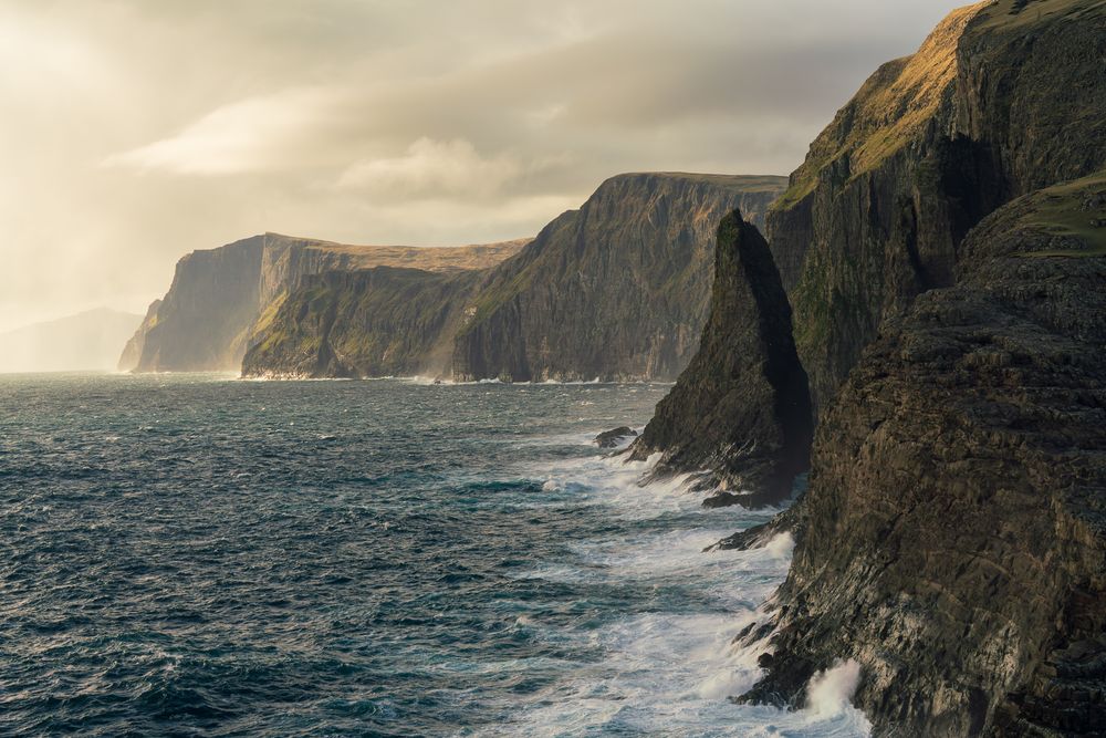 Towering dark sea cliffs rise above turbulent ocean waves under a moody stormy sky.