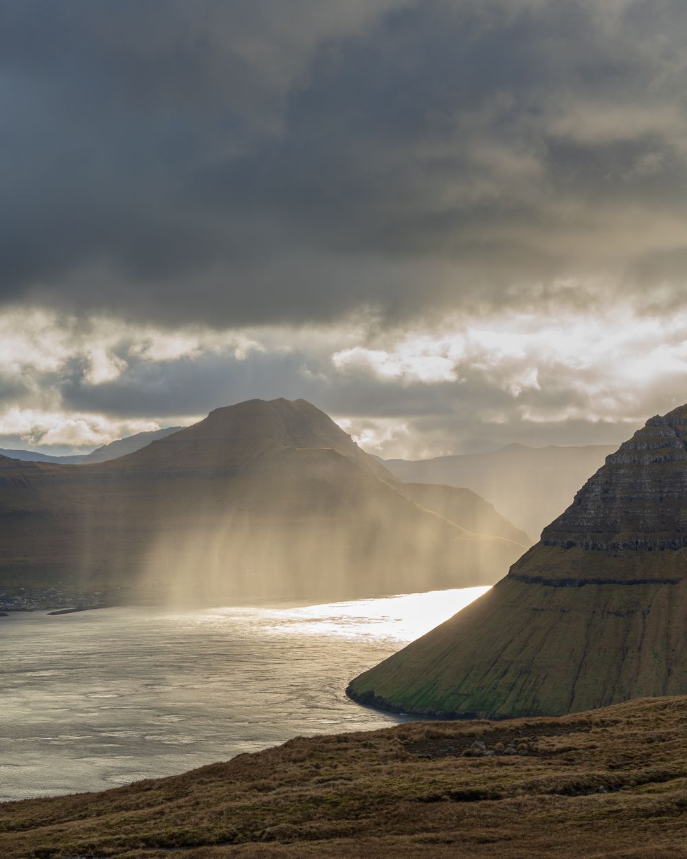 Dramatic rays of light pierce storm clouds over a misty fjord flanked by steep mountains.