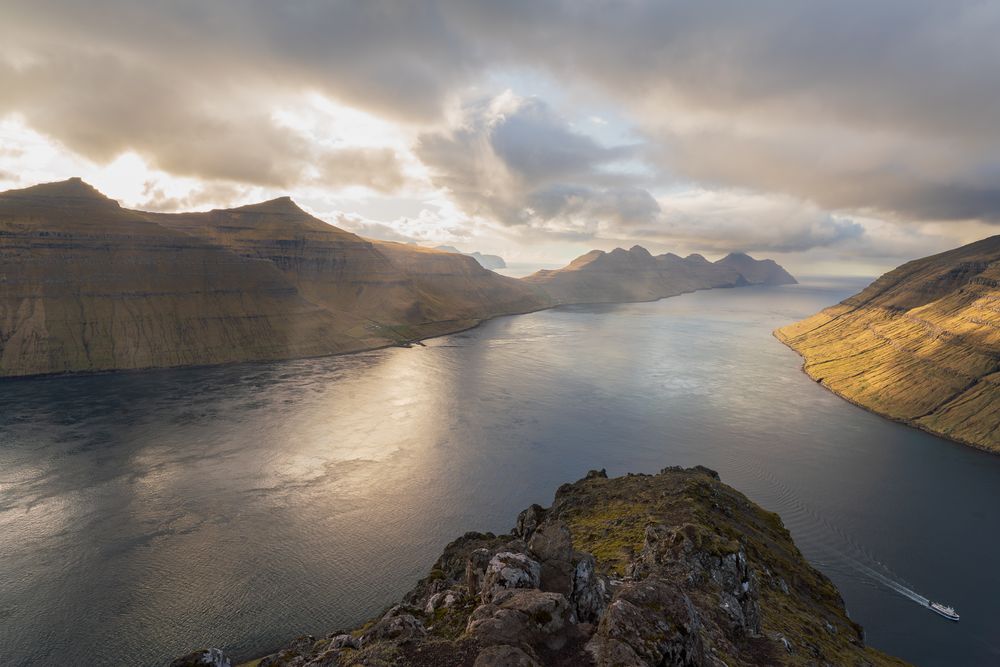 Golden sunlight breaks through dramatic clouds over a serene fjord surrounded by mountains.