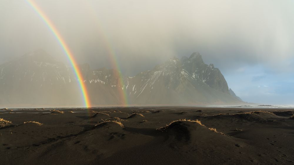 Double rainbow over black sand dunes with dramatic misty mountains in the background in Iceland.