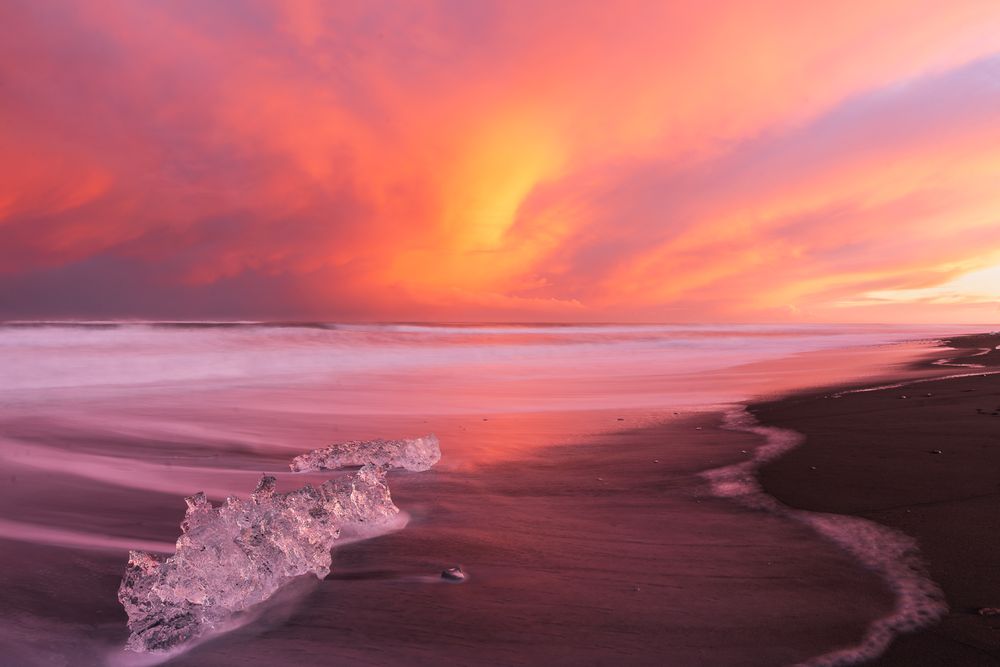 A vivid pink and orange sunset illuminates the sky above a black sand beach with ice chunk.