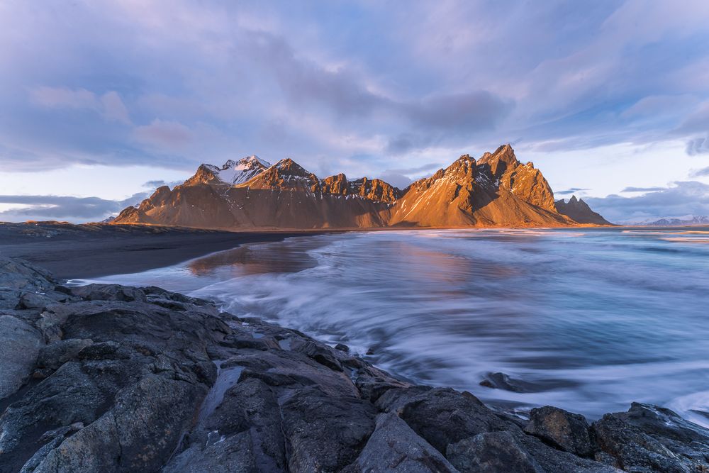 Golden sunset light on Vestrahorn mountain peaks reflecting over black sand beach in Iceland.