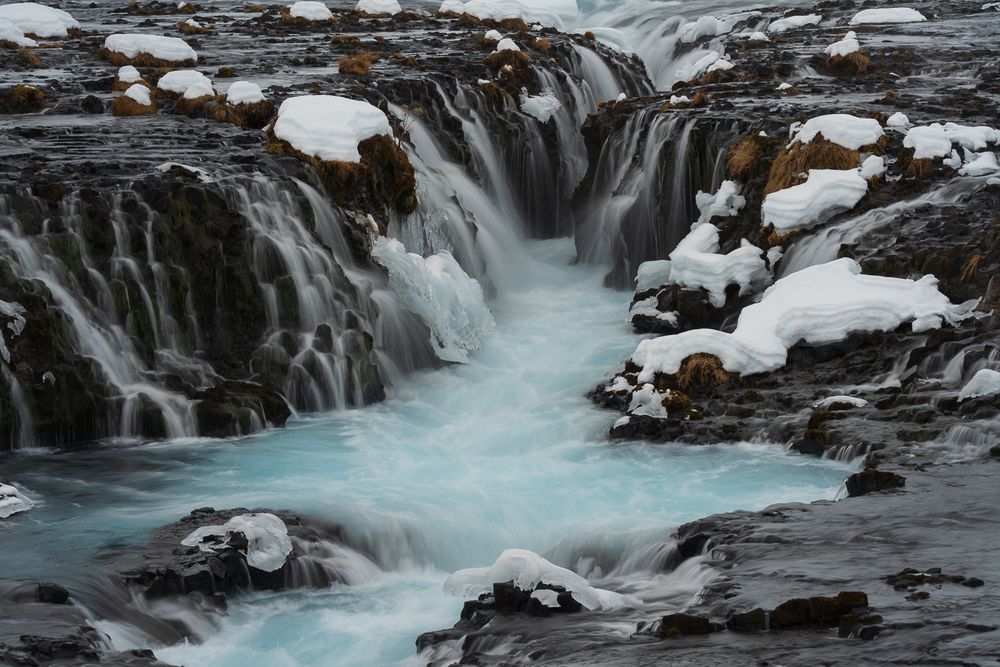 Turquoise glacial water rushing through snow-covered rocks at Bruarfoss waterfall in Iceland.