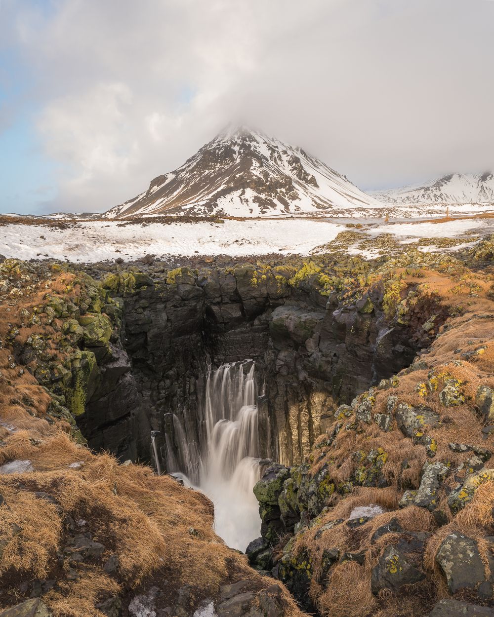 Waterfall cascading into rocky gorge with snow-capped volcanic mountain in Iceland winter landscape.