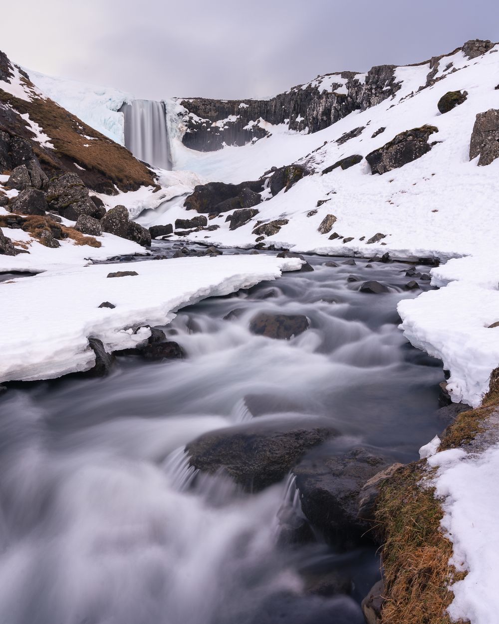 Snowy winter waterfall flowing into icy stream with frozen landscape in Iceland highlands.