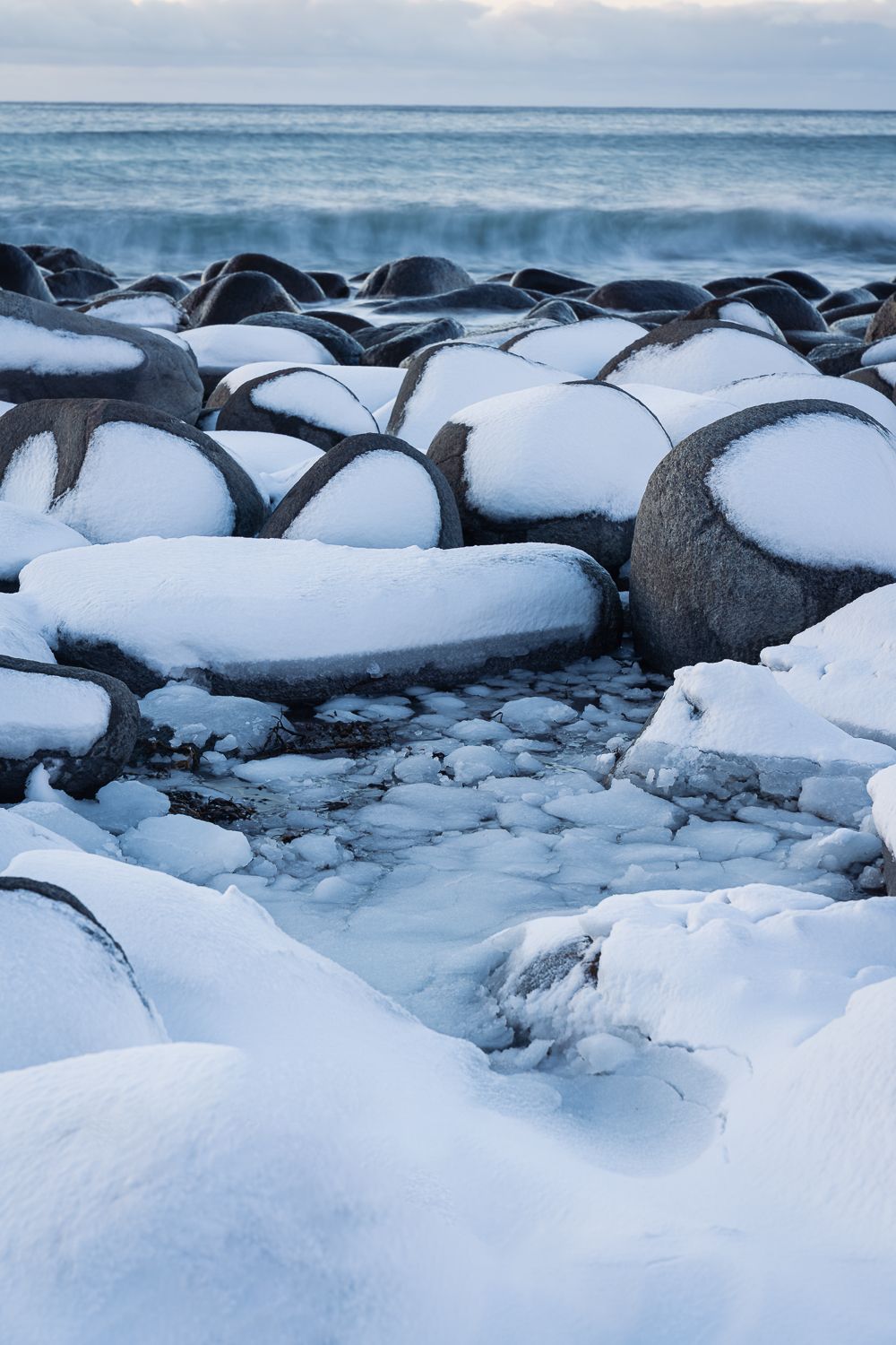Snow-covered large rounded boulders on arctic coastline with ocean waves in winter twilight.