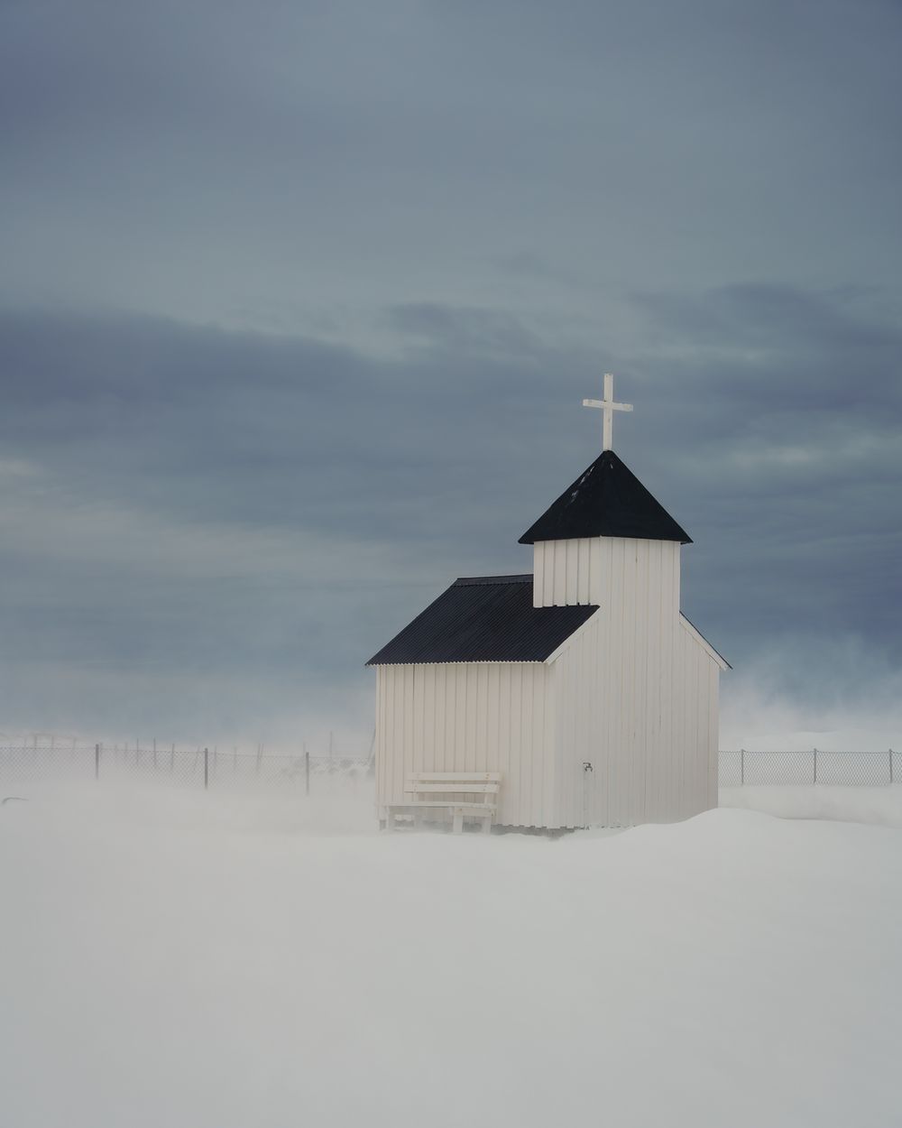 Isolated white wooden church with black roof and cross standing alone in snowy blizzard landscape.