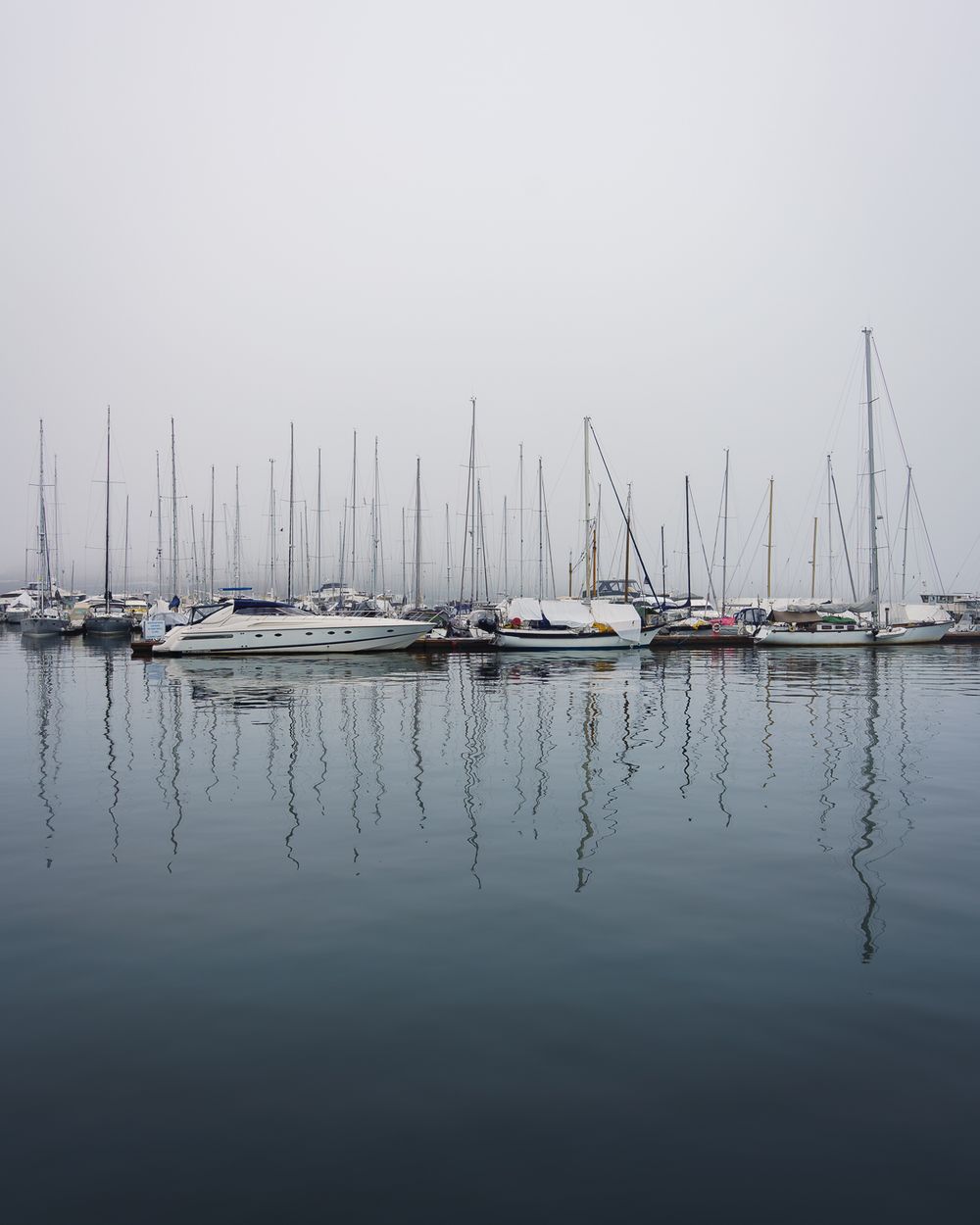 Sailboats moored in calm misty harbor with mast reflections on still grey water.