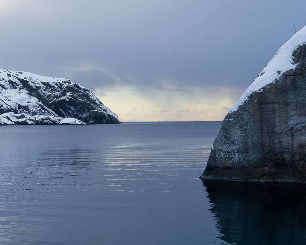 Two snow-capped rocky cliffs frame a calm dark fjord under moody overcast skies in Arctic Norway.