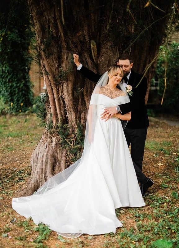 Bride in white gown with veil and groom in black suit pose by a large tree outdoors on their wedding day.