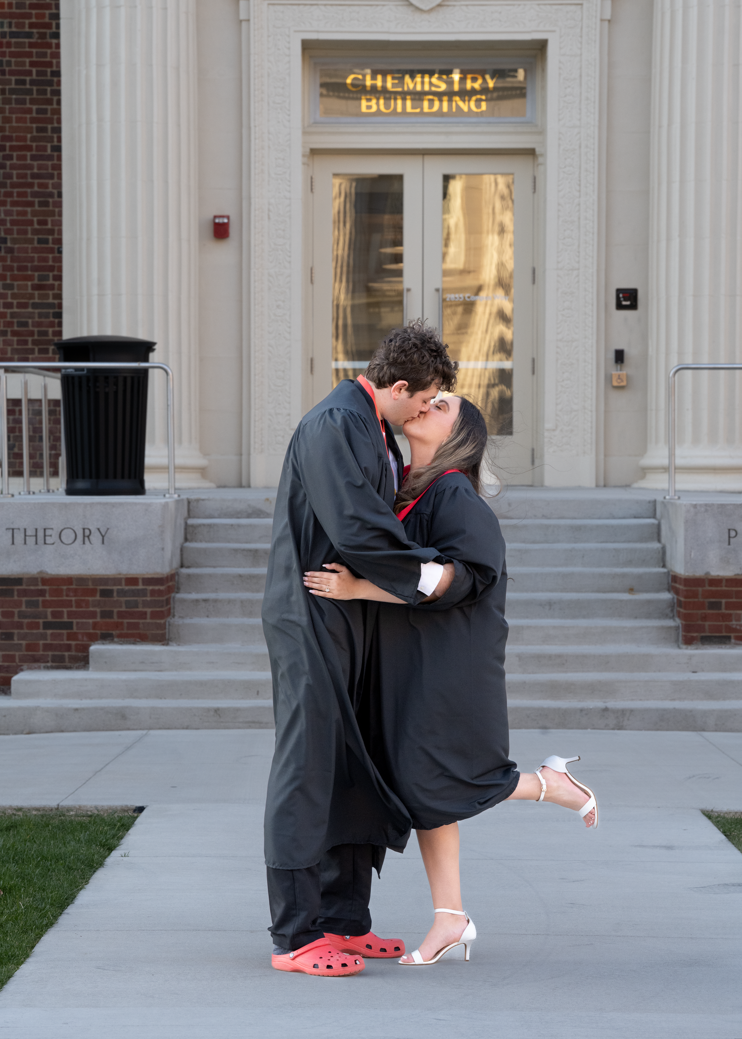 Two graduates in black robes share a romantic kiss outside a Chemistry Building on graduation day.