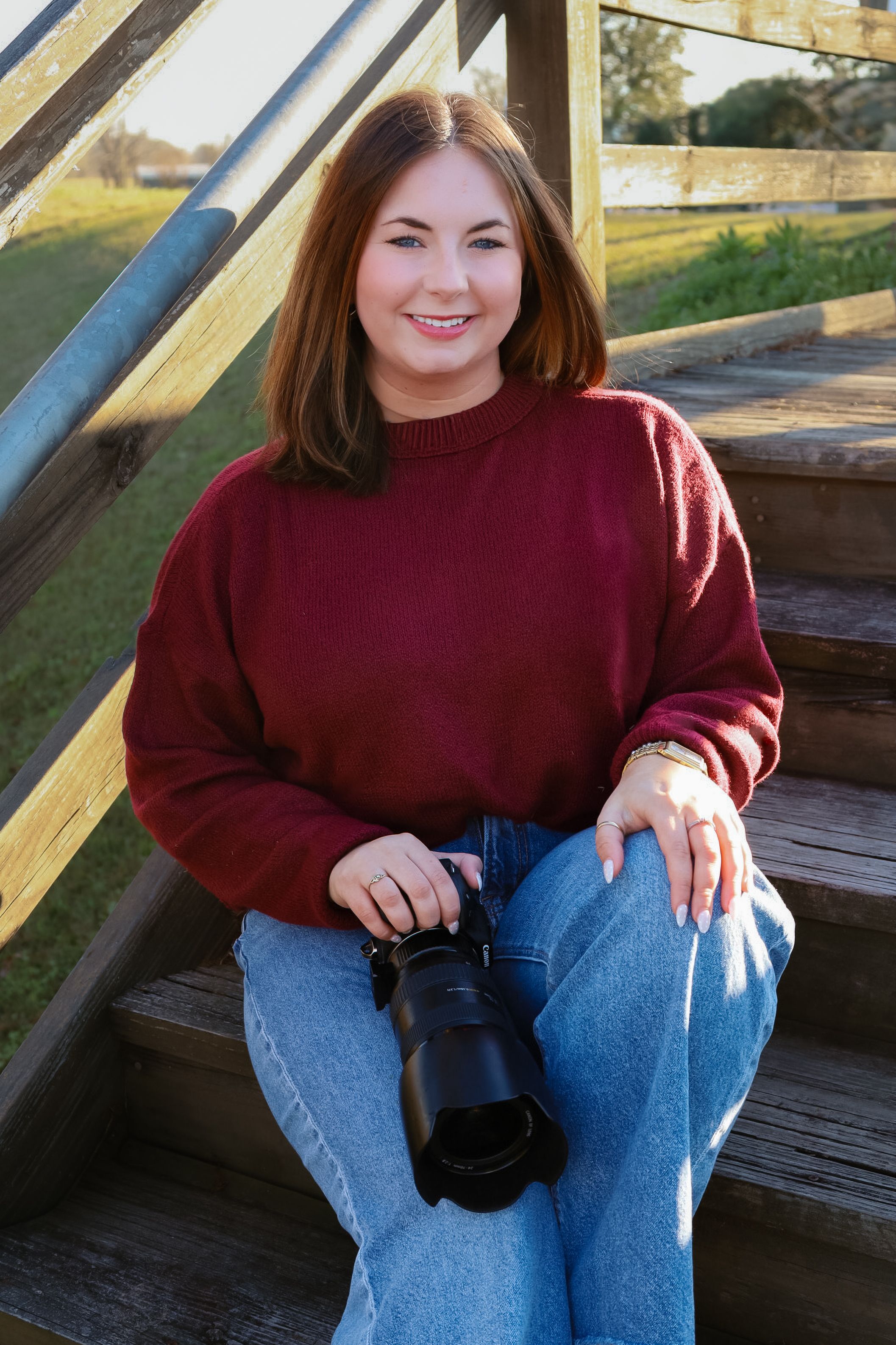 Young woman in red sweater and jeans sits on wooden steps holding a DSLR camera, smiling outdoors.