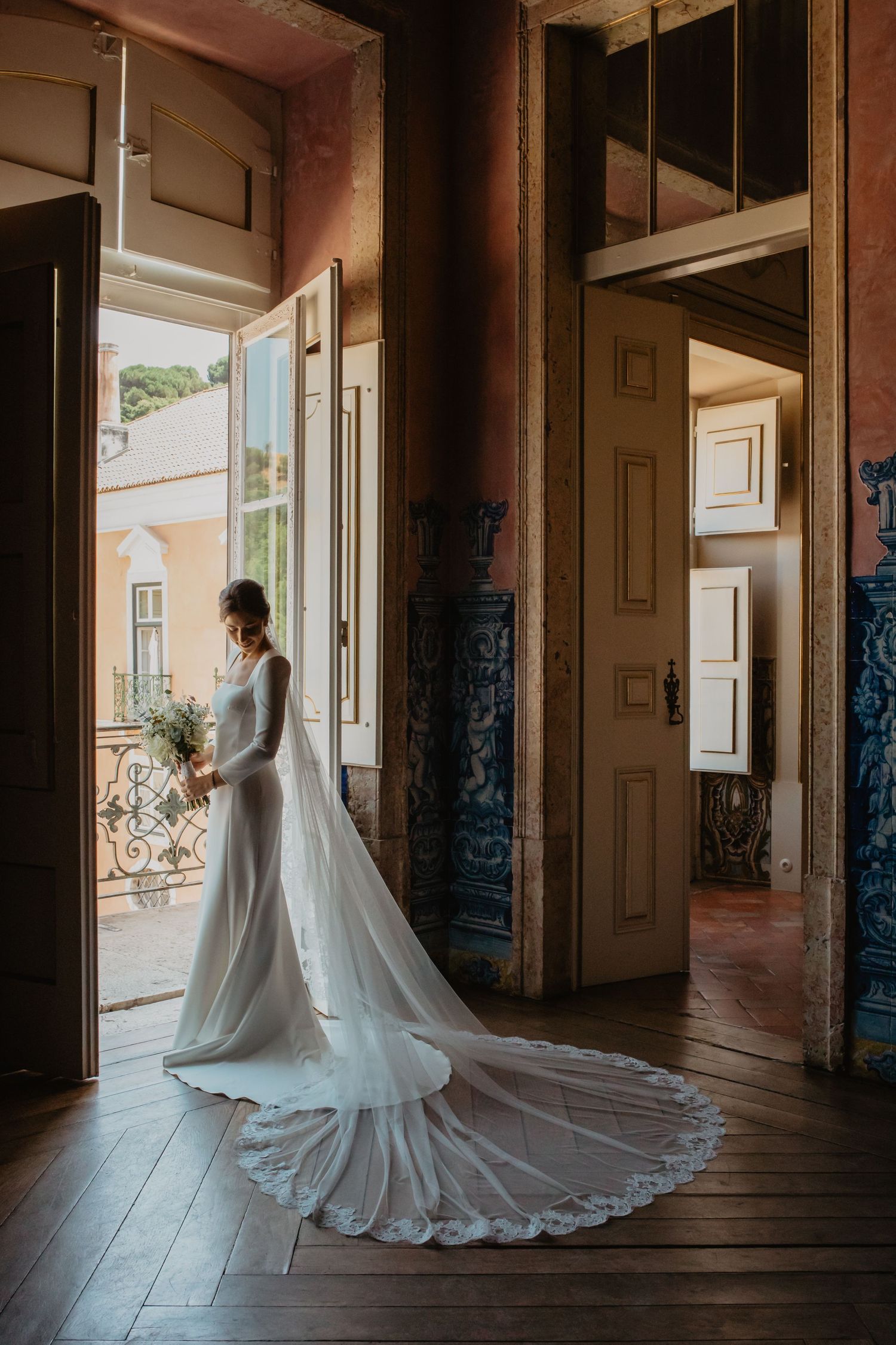 Bride in elegant long-sleeve white gown holding bouquet in ornate tiled hall.