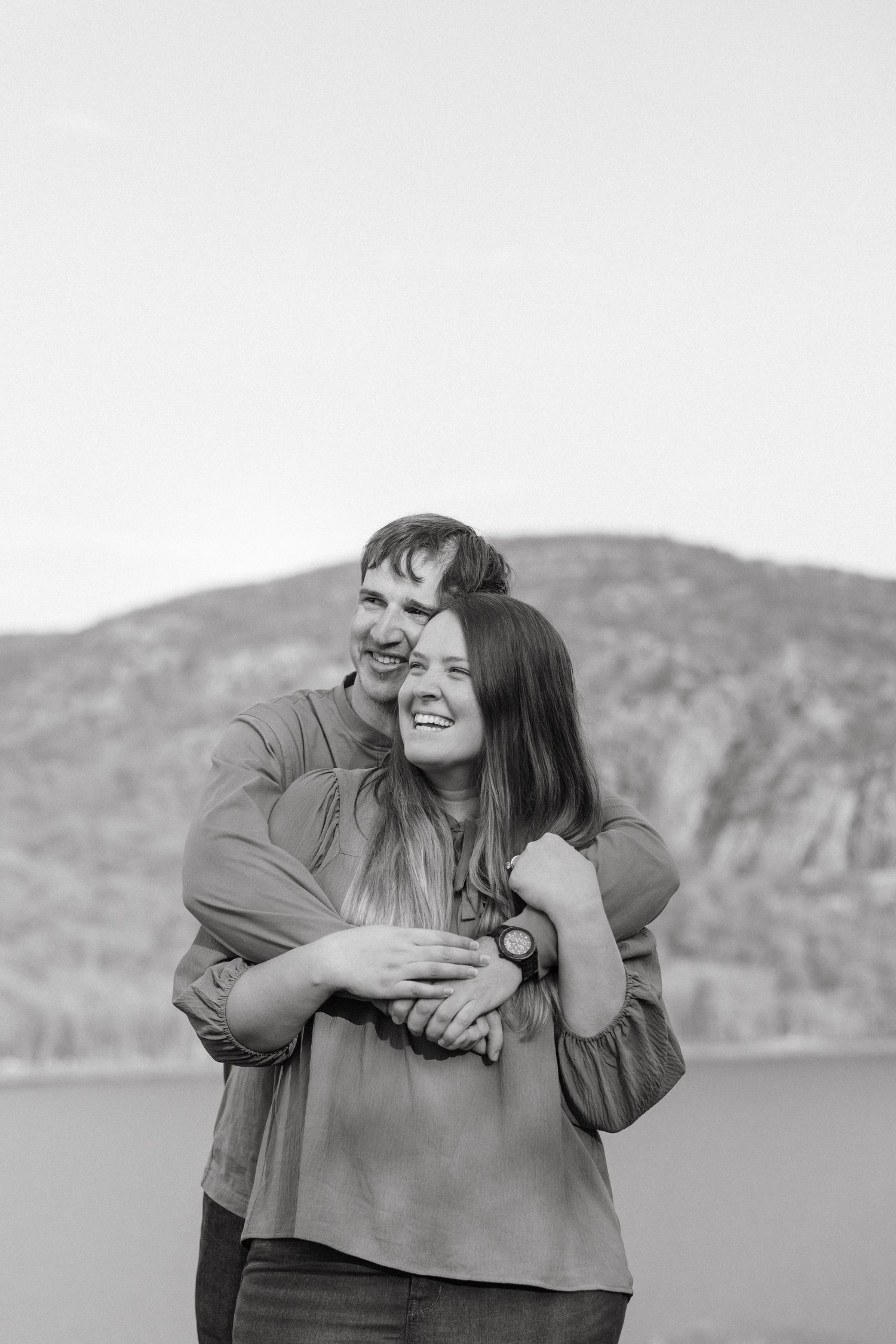 Black and white photo of a laughing couple embracing outdoors in a forest, woman hugging man from behind.