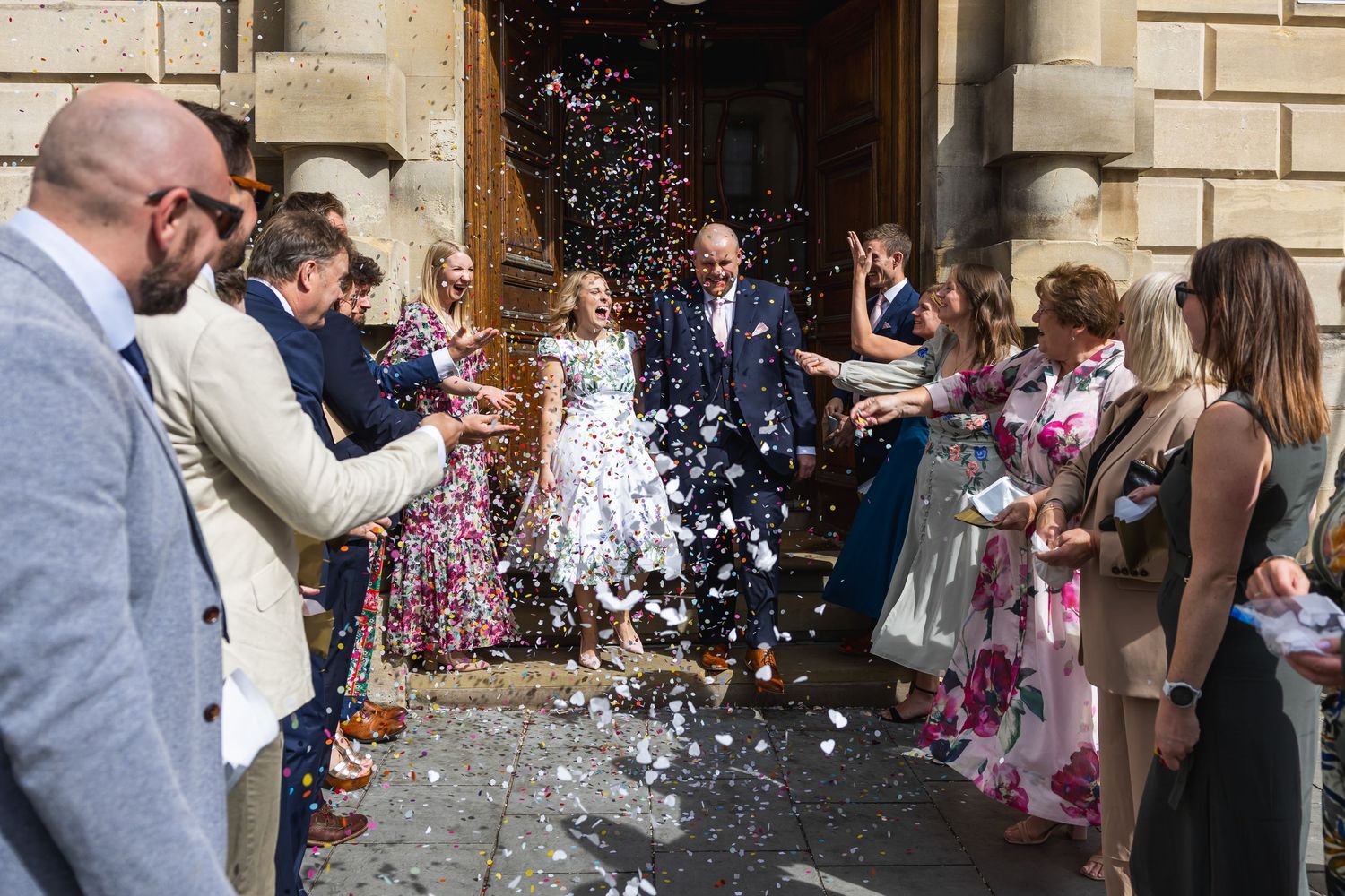 Newlyweds exit Bath's Guildhall amid confetti shower surrounded by cheering wedding guests on sunny day.