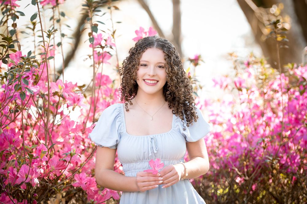Young woman with curly hair smiling in a blue dress surrounded by vibrant pink azalea flowers outdoors.