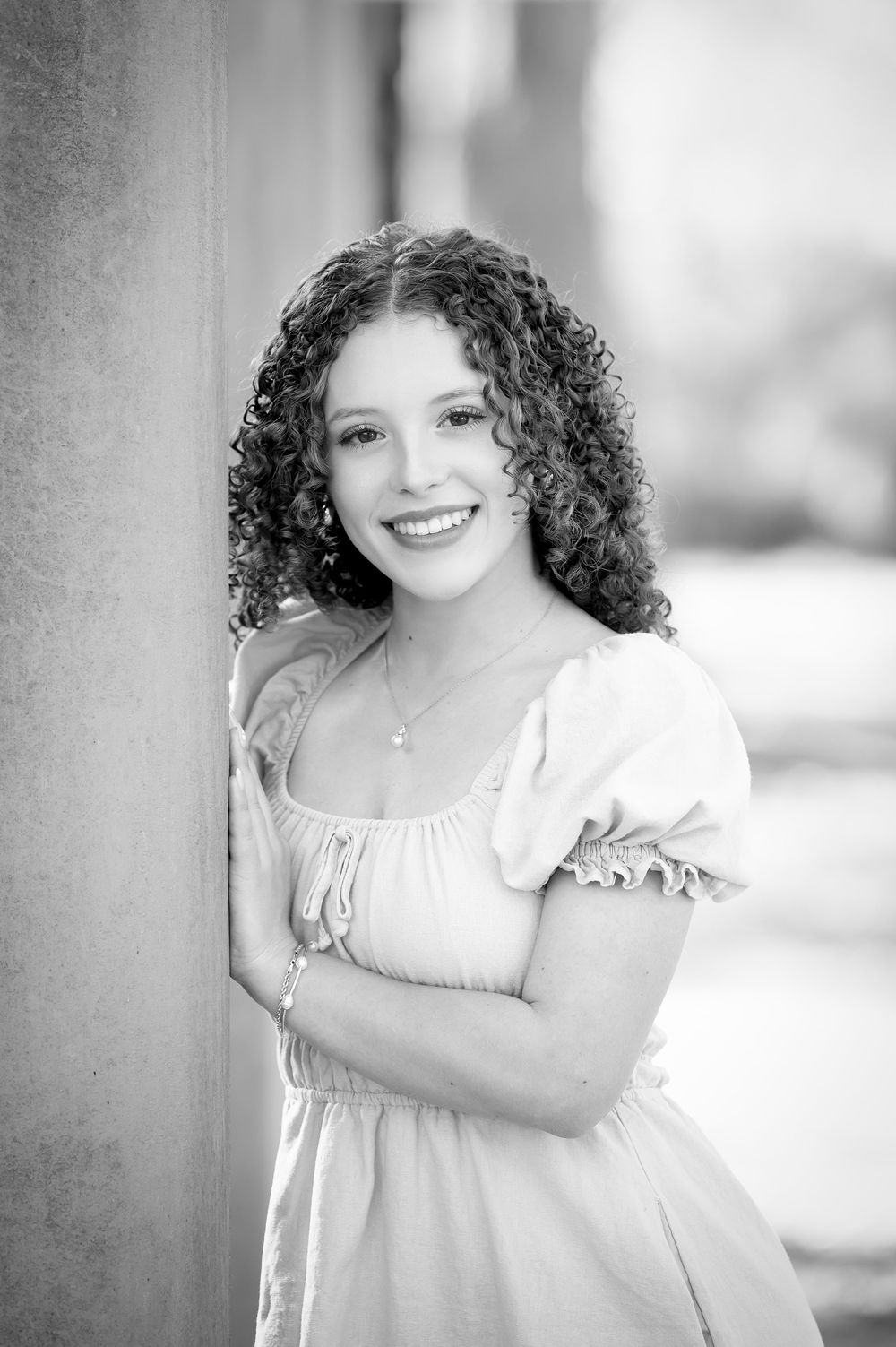 Black and white portrait of a young woman with curly hair in a ruffled dress, smiling warmly while leaning against a column.