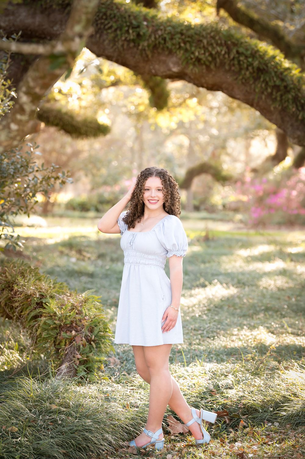 Young woman in white dress poses gracefully under moss-covered oak trees in a sunlit garden with pink blooms.