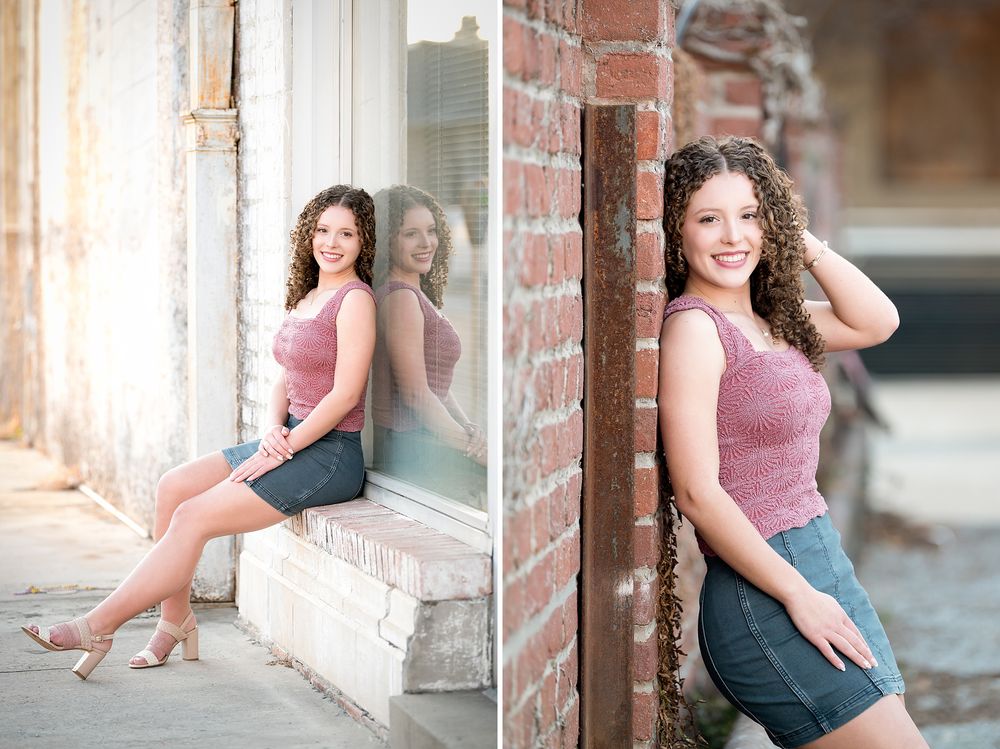 Young woman with curly hair posing in pink tank and denim skirt by brick wall and window outdoors.
