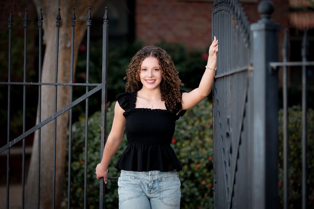 Young woman with curly hair in black top and jeans posing by iron gate and brick wall outdoors.