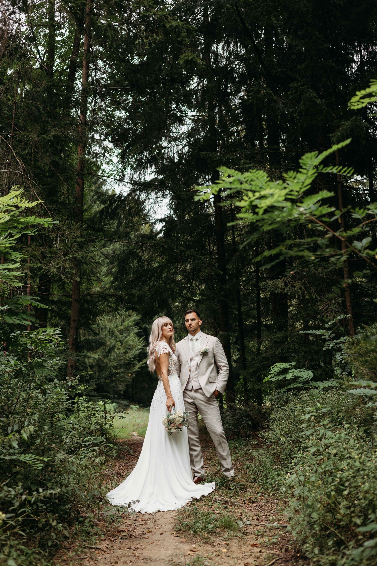 Bride and groom stand on a forest path surrounded by lush green trees in an elegant outdoor wedding photo.