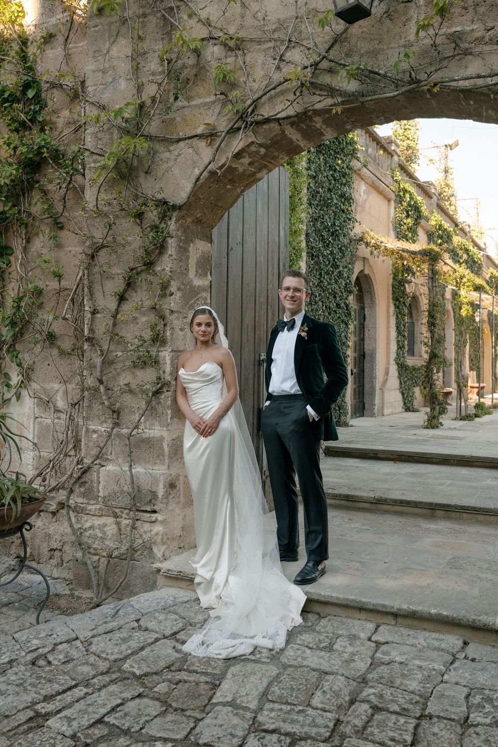 Bride and groom posing beneath ivy-draped stone arch on rustic cobblestone courtyard in wedding attire.