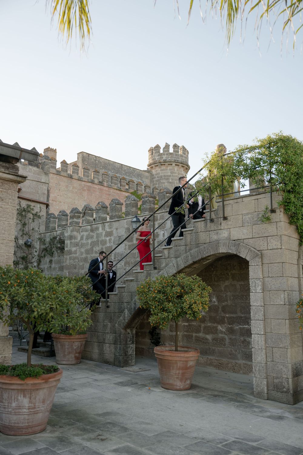 Wedding guests descend stone castle staircase with potted citrus trees and medieval battlements.