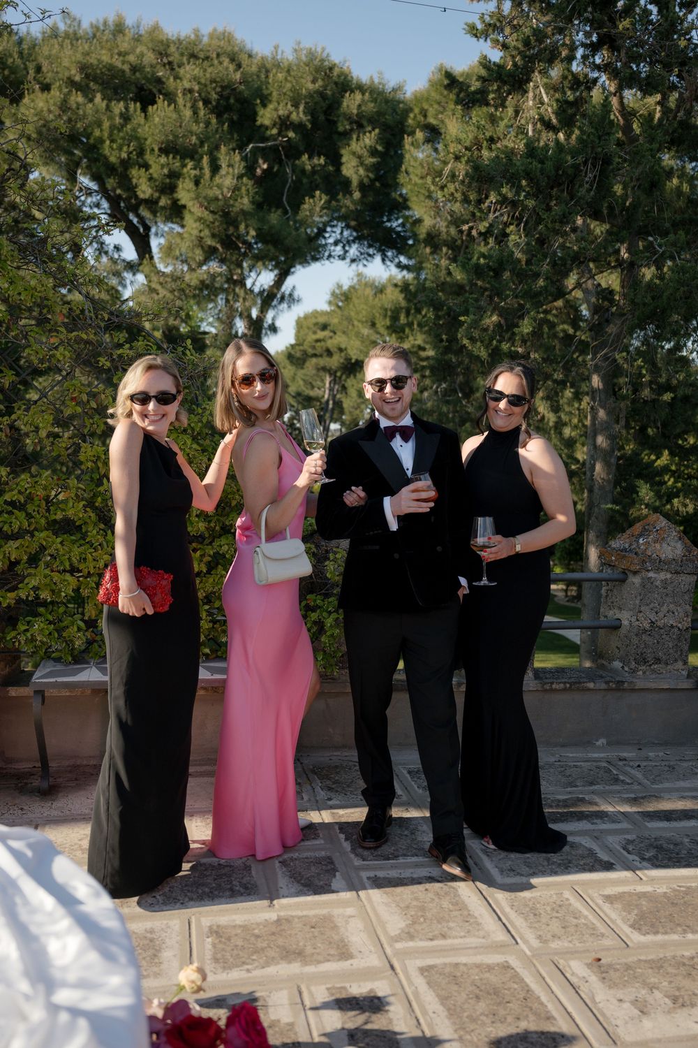 Four elegantly dressed wedding guests pose outdoors, three in black gowns and one in pink, holding champagne glasses.
