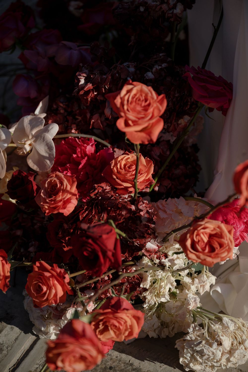 Lush wedding floral arrangement featuring coral roses, deep red dahlias, and white orchids.