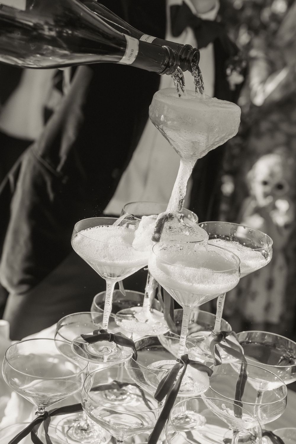 Black and white photo of champagne being poured into a pyramid tower of coupe glasses at an elegant celebration.