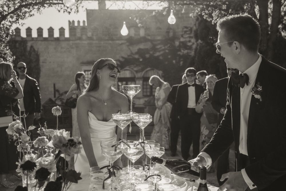 Black and white photo of bride and groom smiling beside a champagne tower at an elegant outdoor wedding reception.