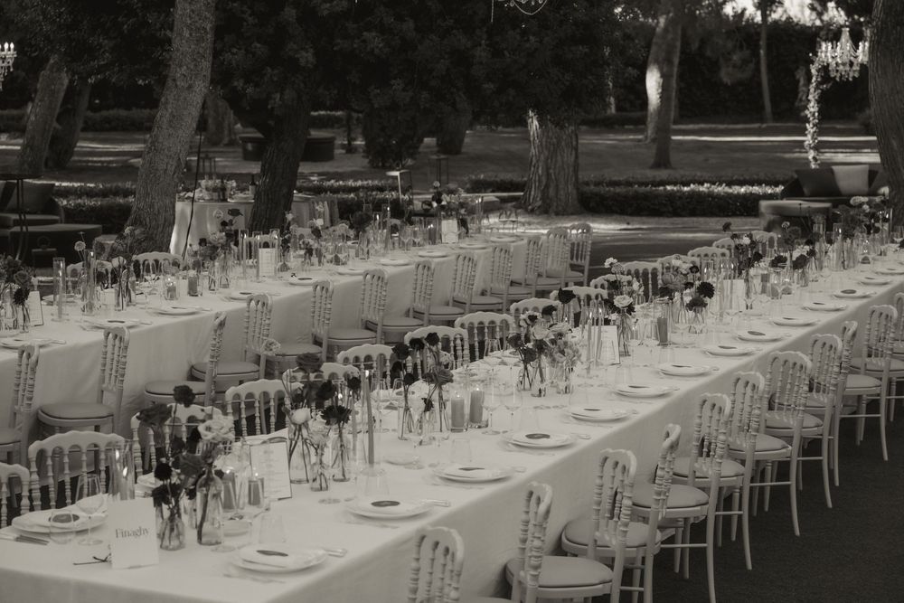 Elegant outdoor wedding reception with long white-draped tables, floral centerpieces, and ornate chairs in a garden setting.