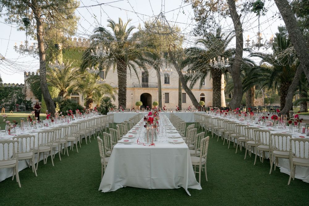 Elegant outdoor wedding reception with long white tables, chiavari chairs, floral centerpieces, and chandeliers set before a historic villa.