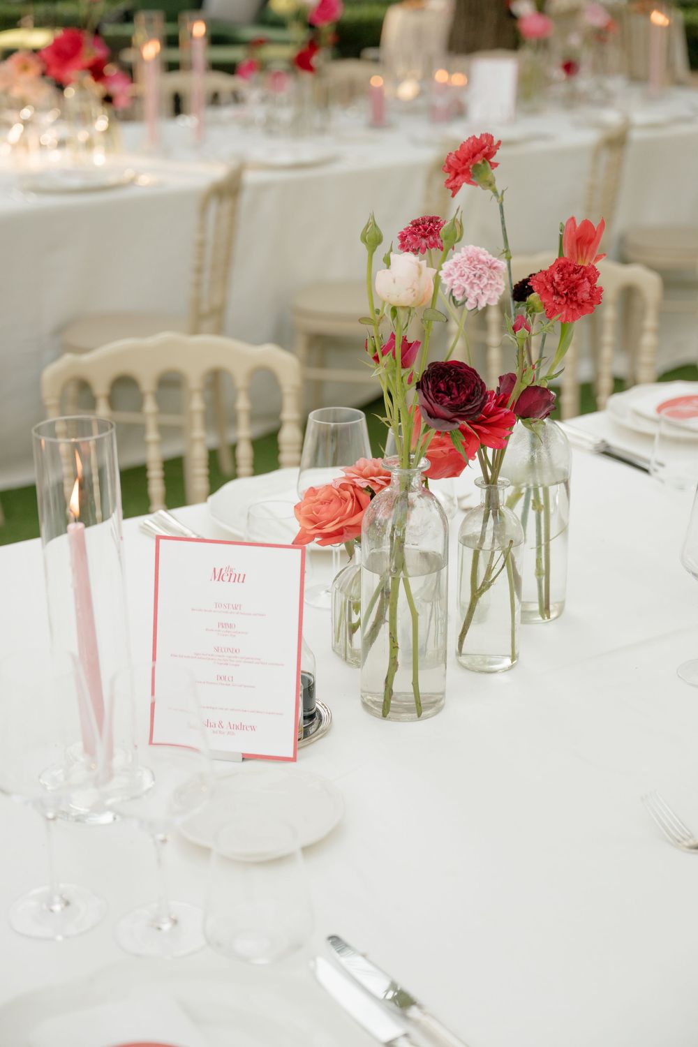 Elegant wedding reception table with pink and red floral arrangements in glass bottles and menu cards.