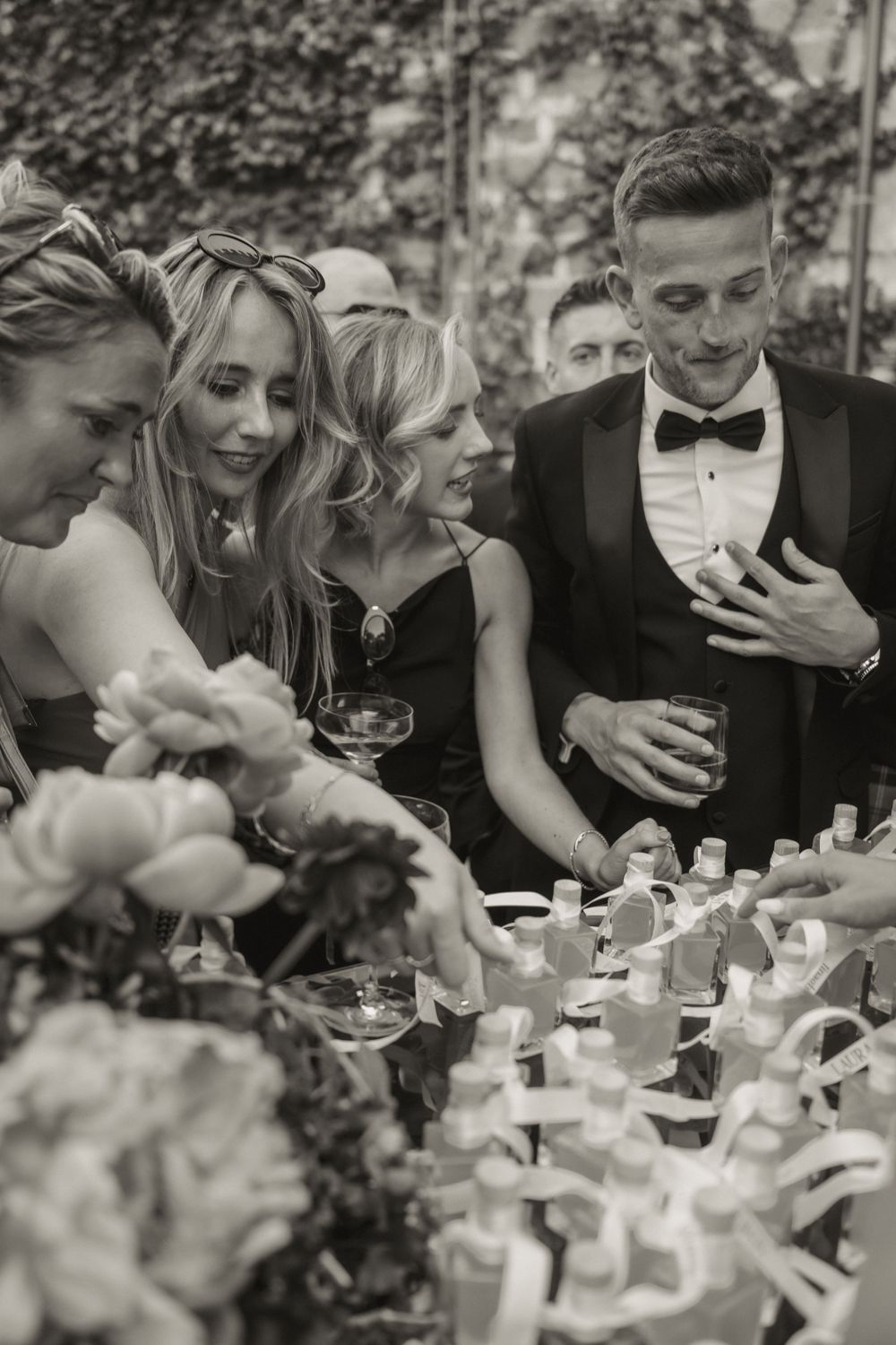 Wedding guests in formal attire gather around a decorated table with floral arrangements in black and white photo.