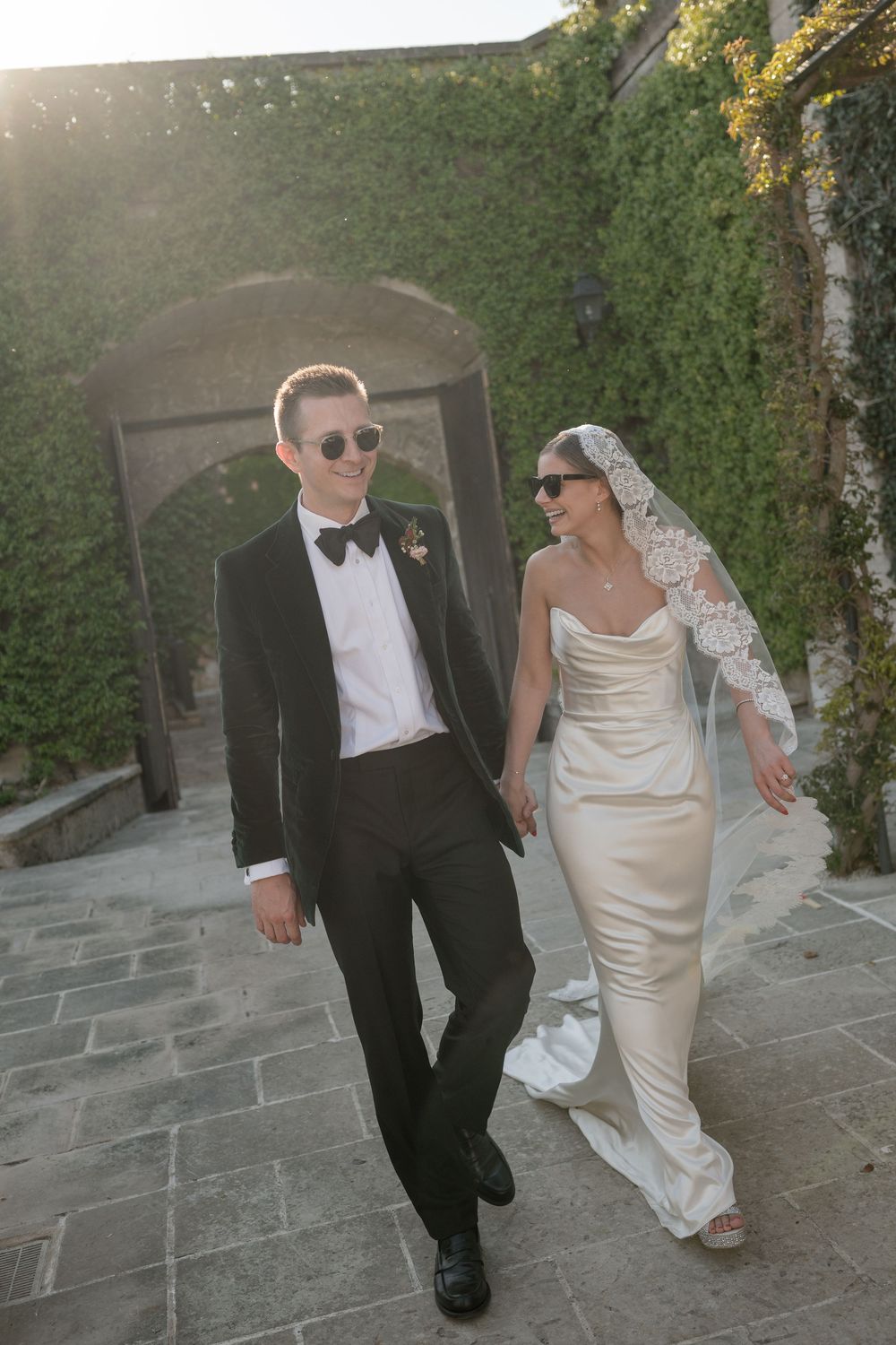 Bride in satin gown and groom in black tuxedo walking together outdoors at a sunlit wedding venue.