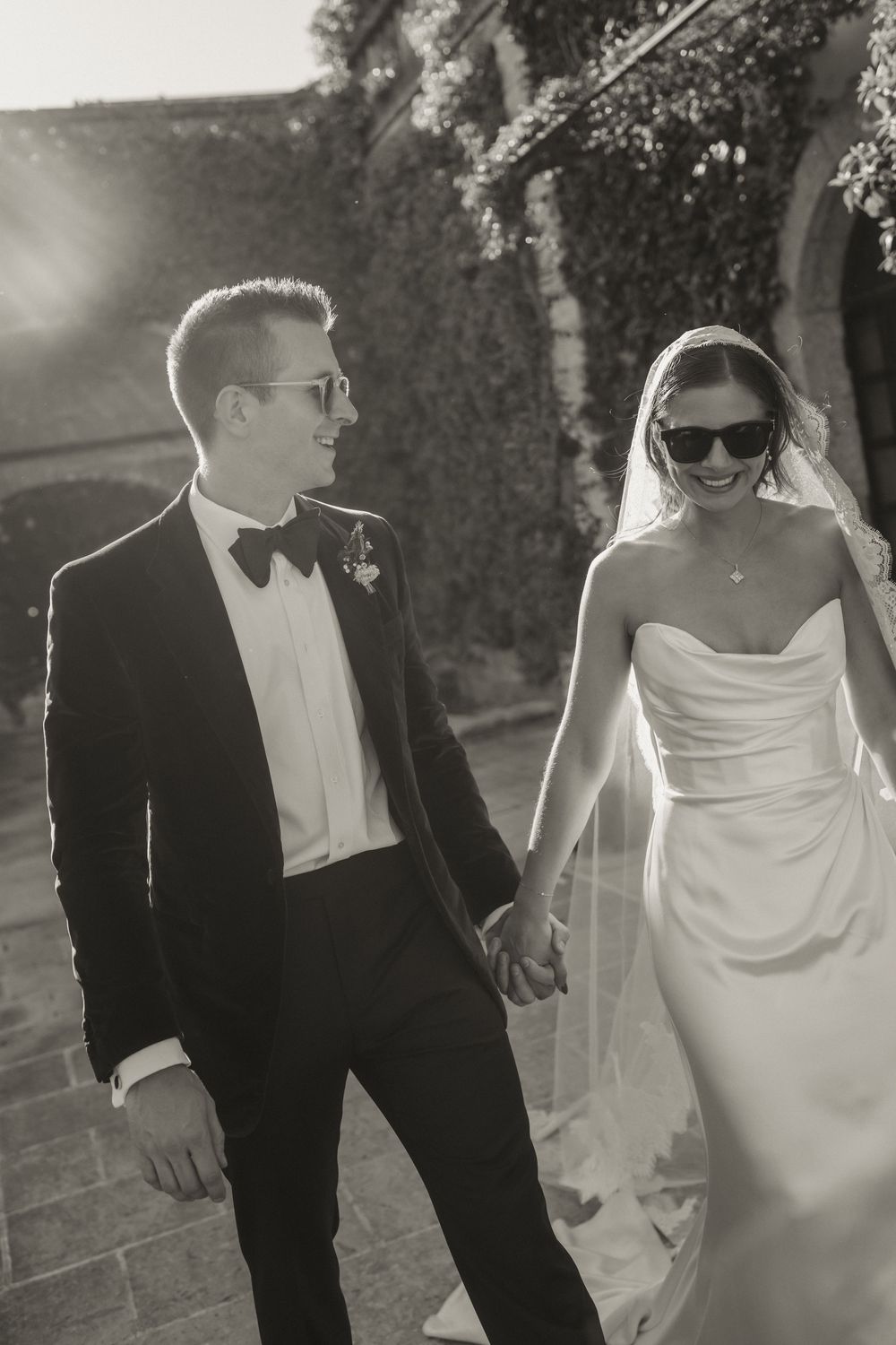 Black and white photo of smiling bride and groom in sunglasses holding hands on their wedding day.