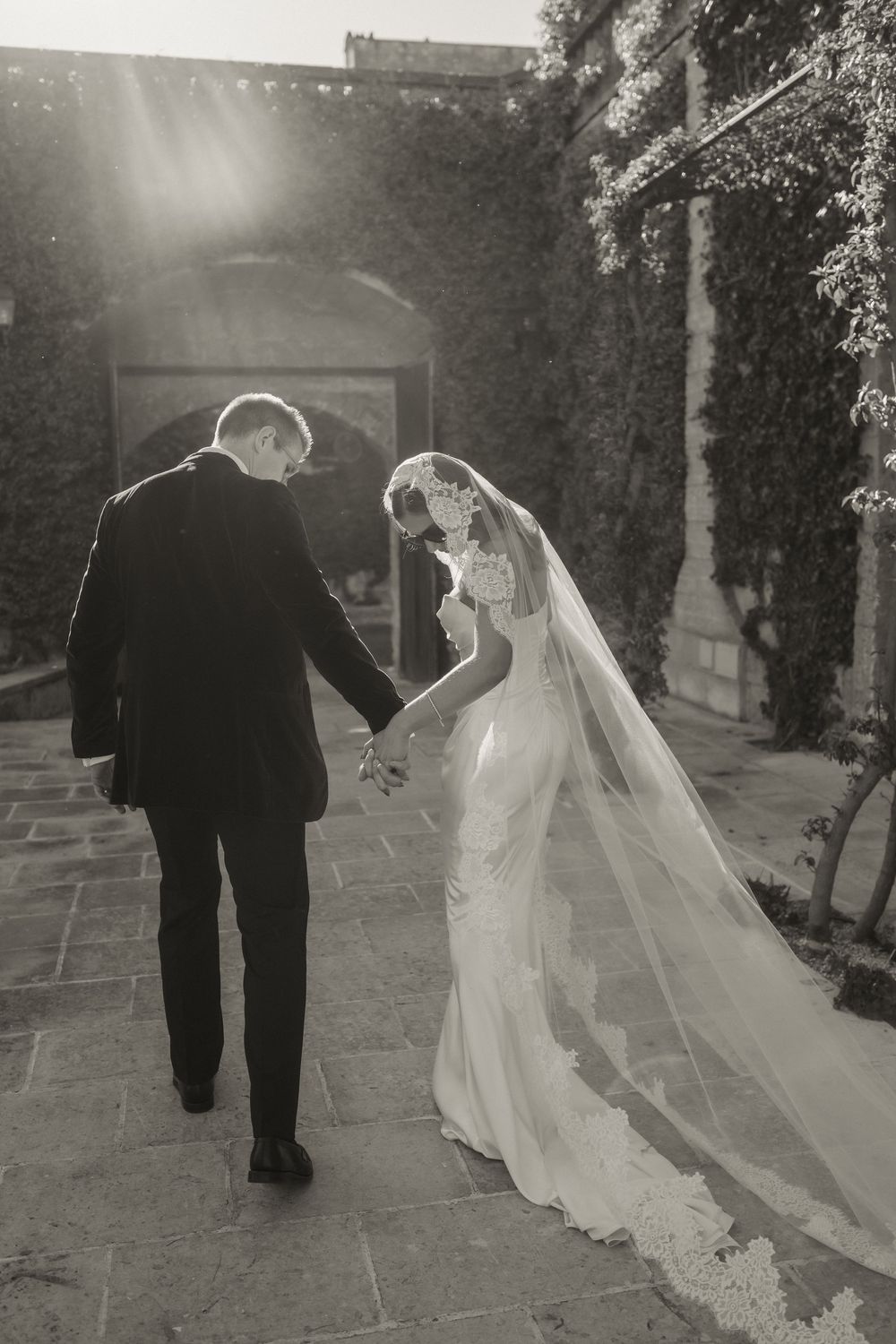 Black and white photo of bride and groom holding hands walking toward arched ivy-covered wedding venue.