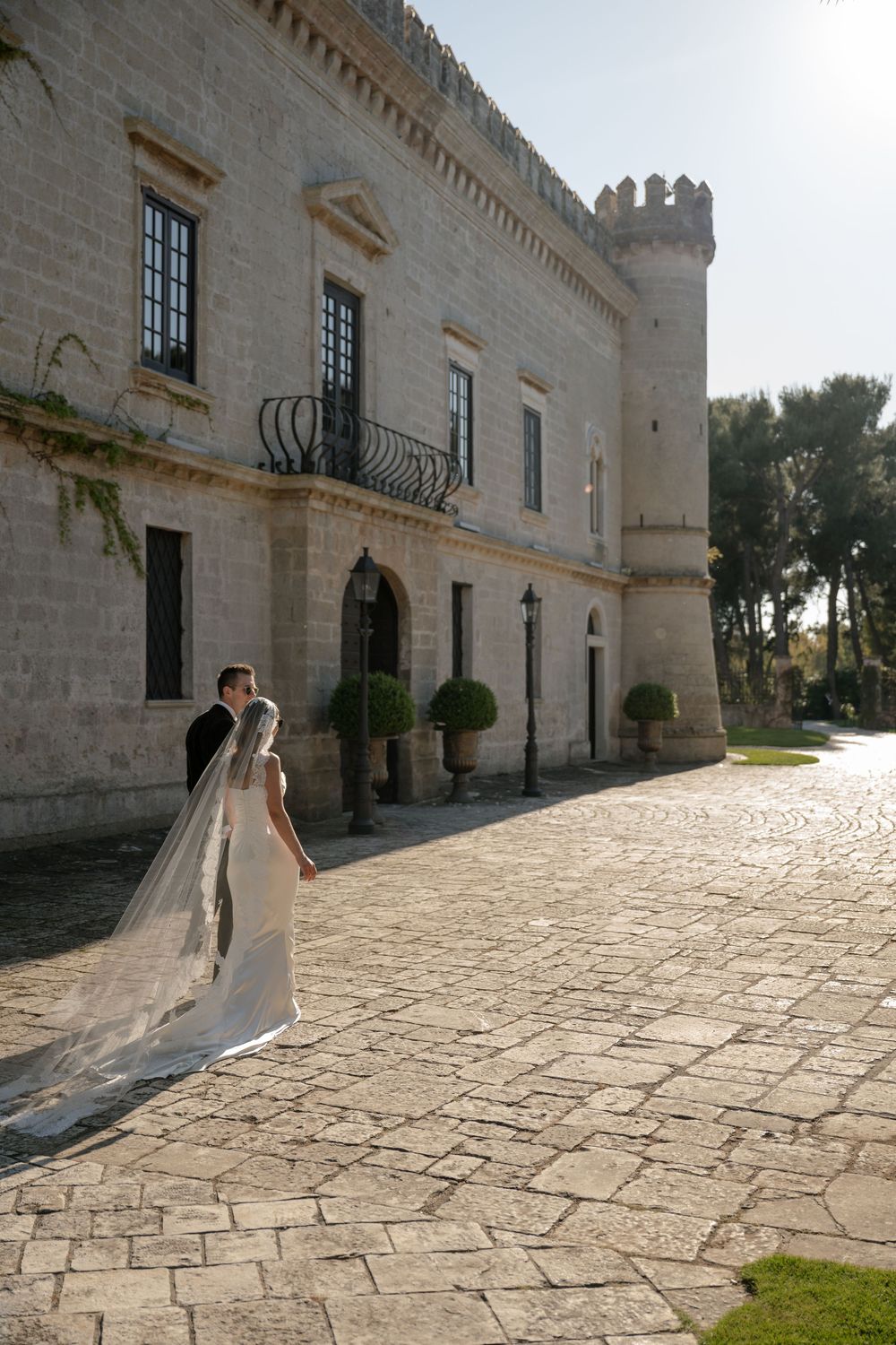 Bride and groom stand before a stunning historic stone castle on their wedding day, bride's long veil trailing on cobblestone.