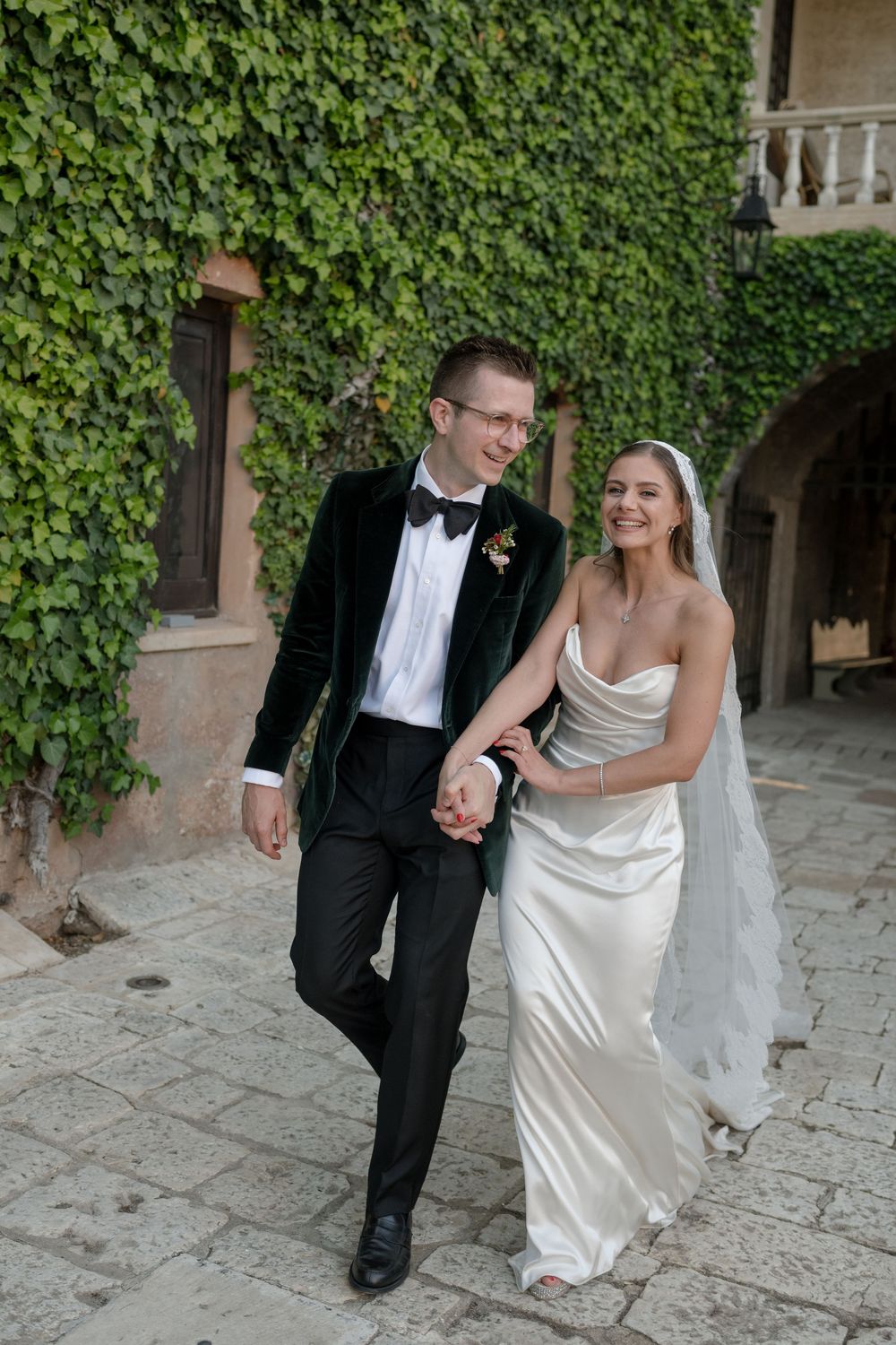Bride and groom walk hand in hand on cobblestone path with ivy-covered walls at elegant wedding venue.