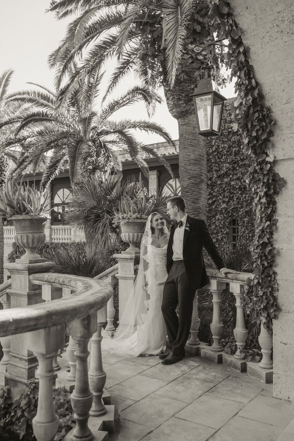 Black and white photo of bride and groom laughing on ornate balcony with palm trees and ivy-covered walls.