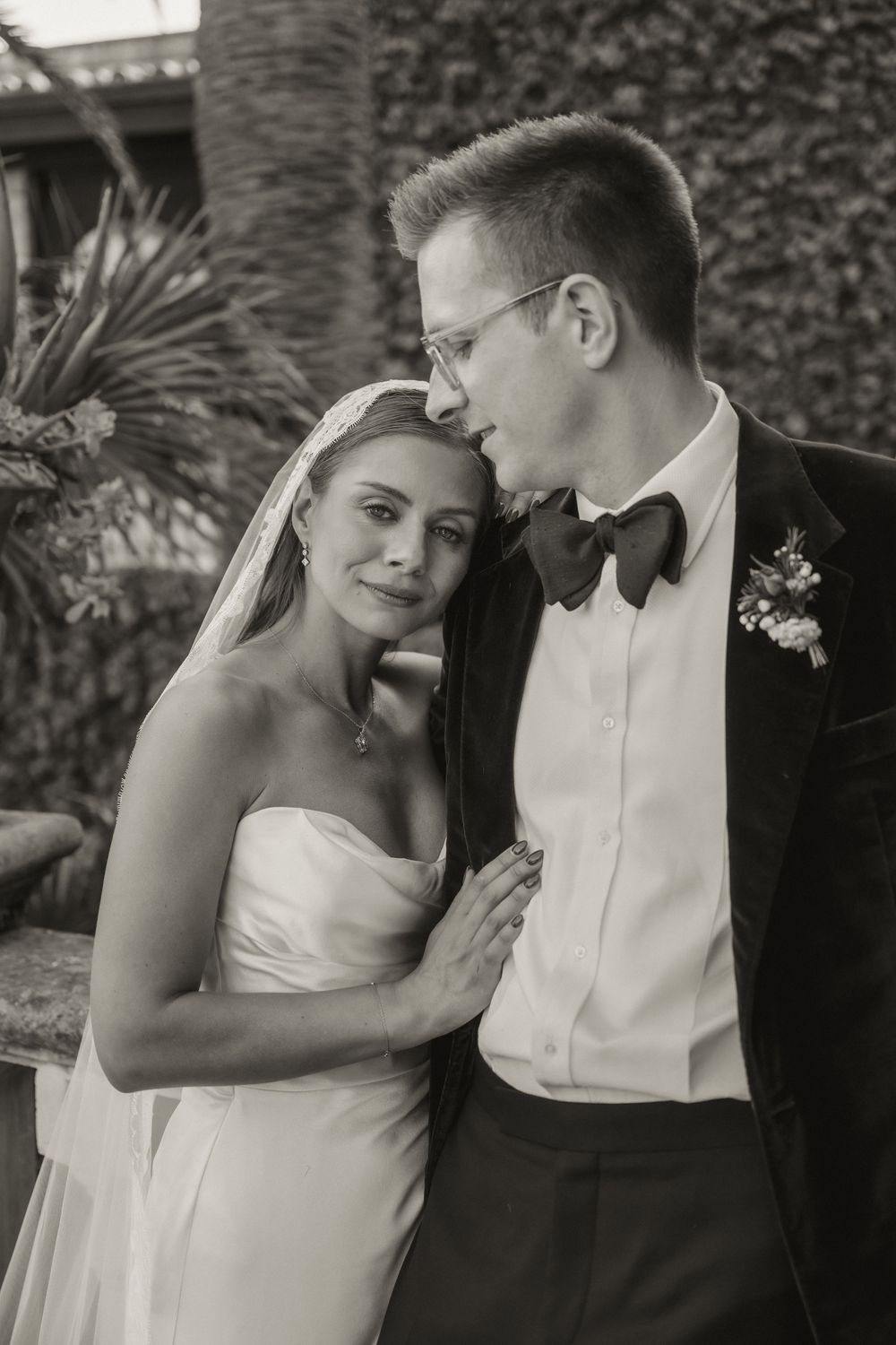 Black and white close-up of groom kissing bride's forehead as she leans against him in wedding attire.