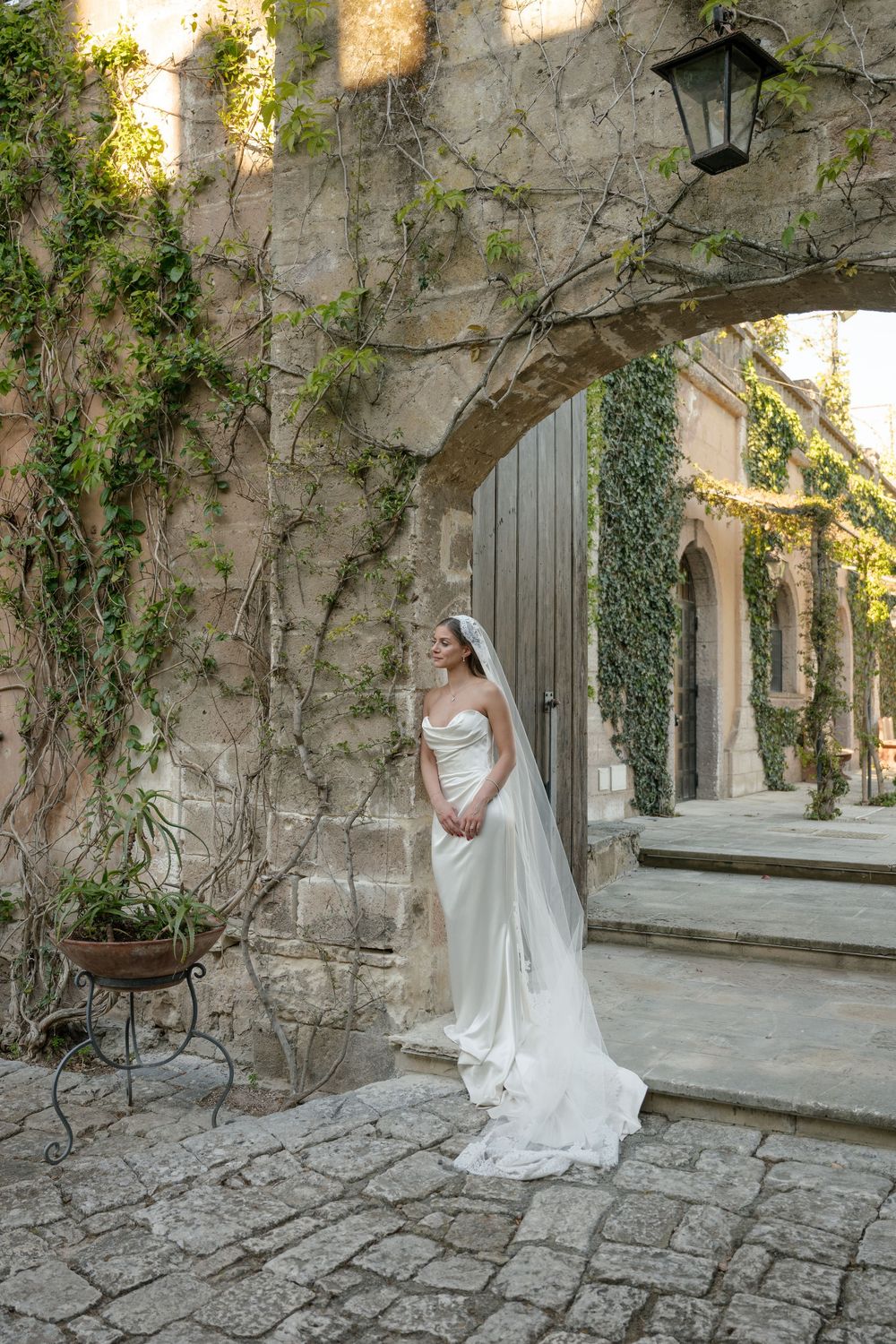Elegant bride in strapless gown with long veil standing beneath ivy-covered stone arch on cobblestone courtyard.