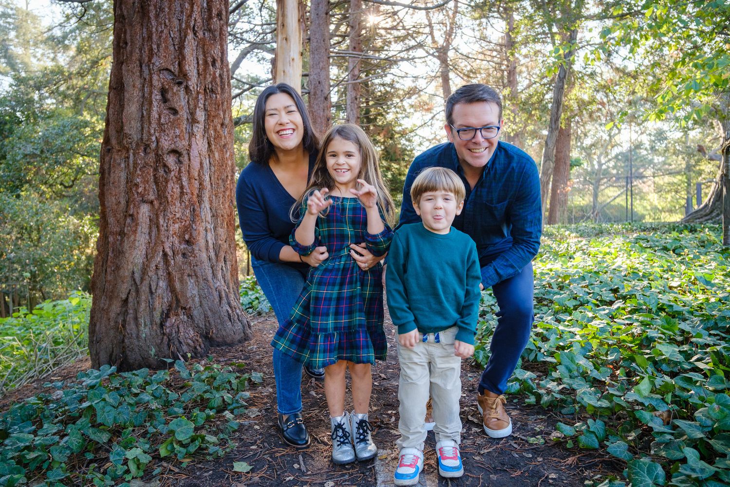 Happy family of four posing outdoors among tall redwood trees in a lush green forest setting.