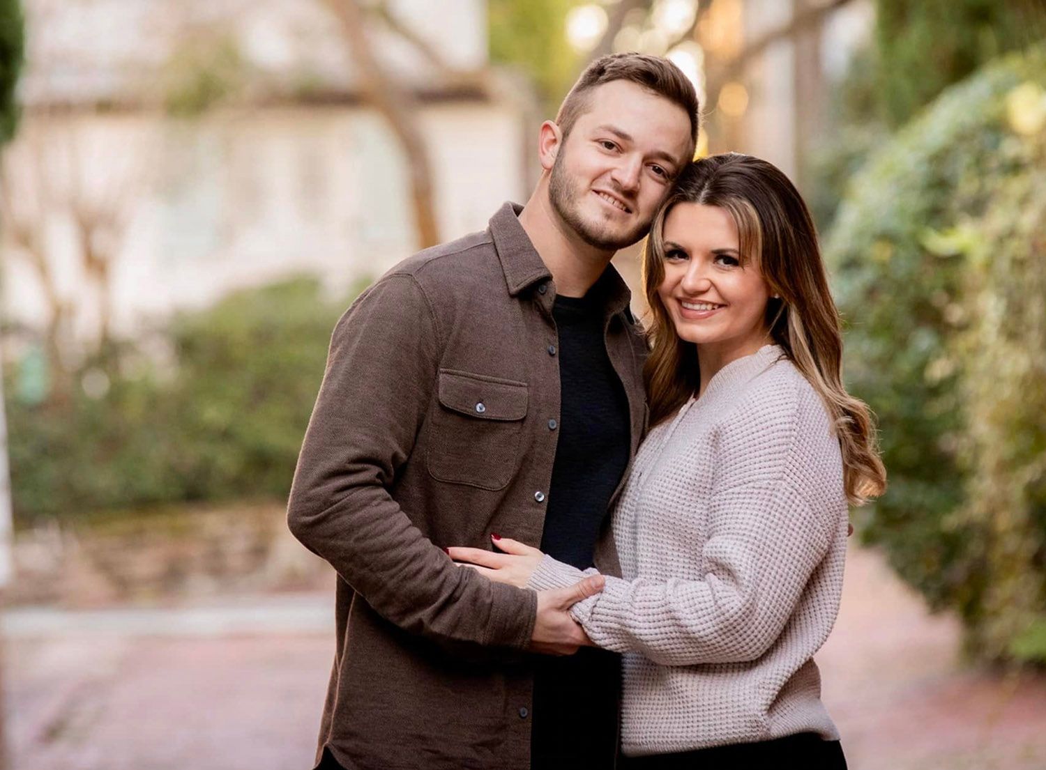A smiling couple poses outdoors, the man in a brown jacket embracing a woman in a beige sweater, with greenery behind them.