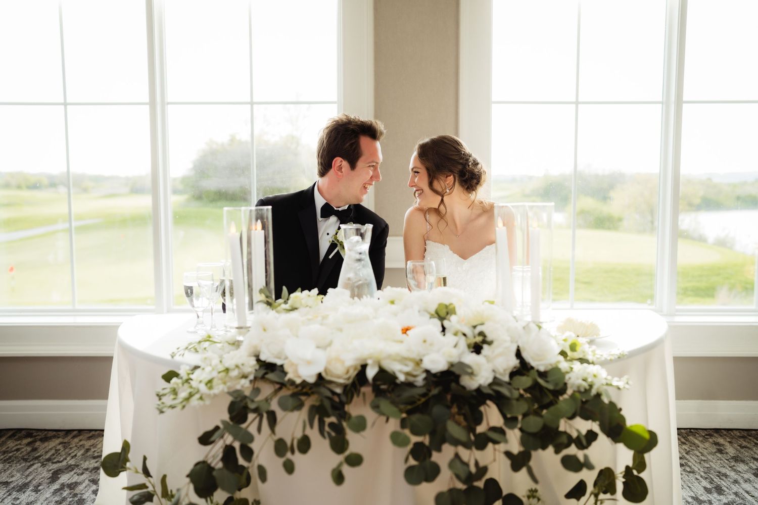 Bride and groom share an intimate moment at floral-adorned sweetheart table with white roses and greenery.
