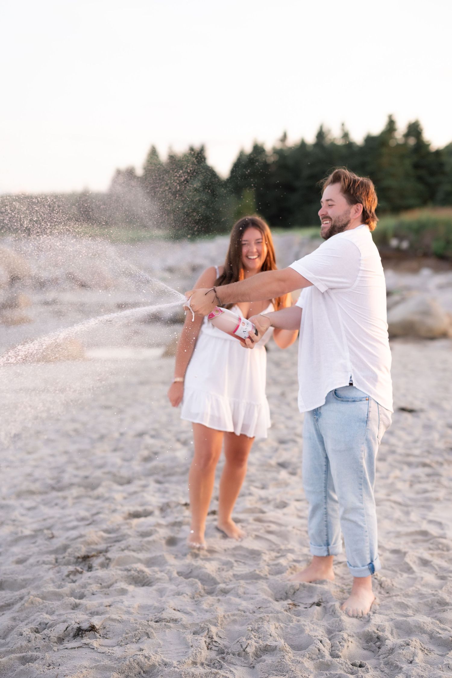 Newly engaged couple popping champagne at golden hour in coastal Nova Scotia to celebrate a surprise proposal.