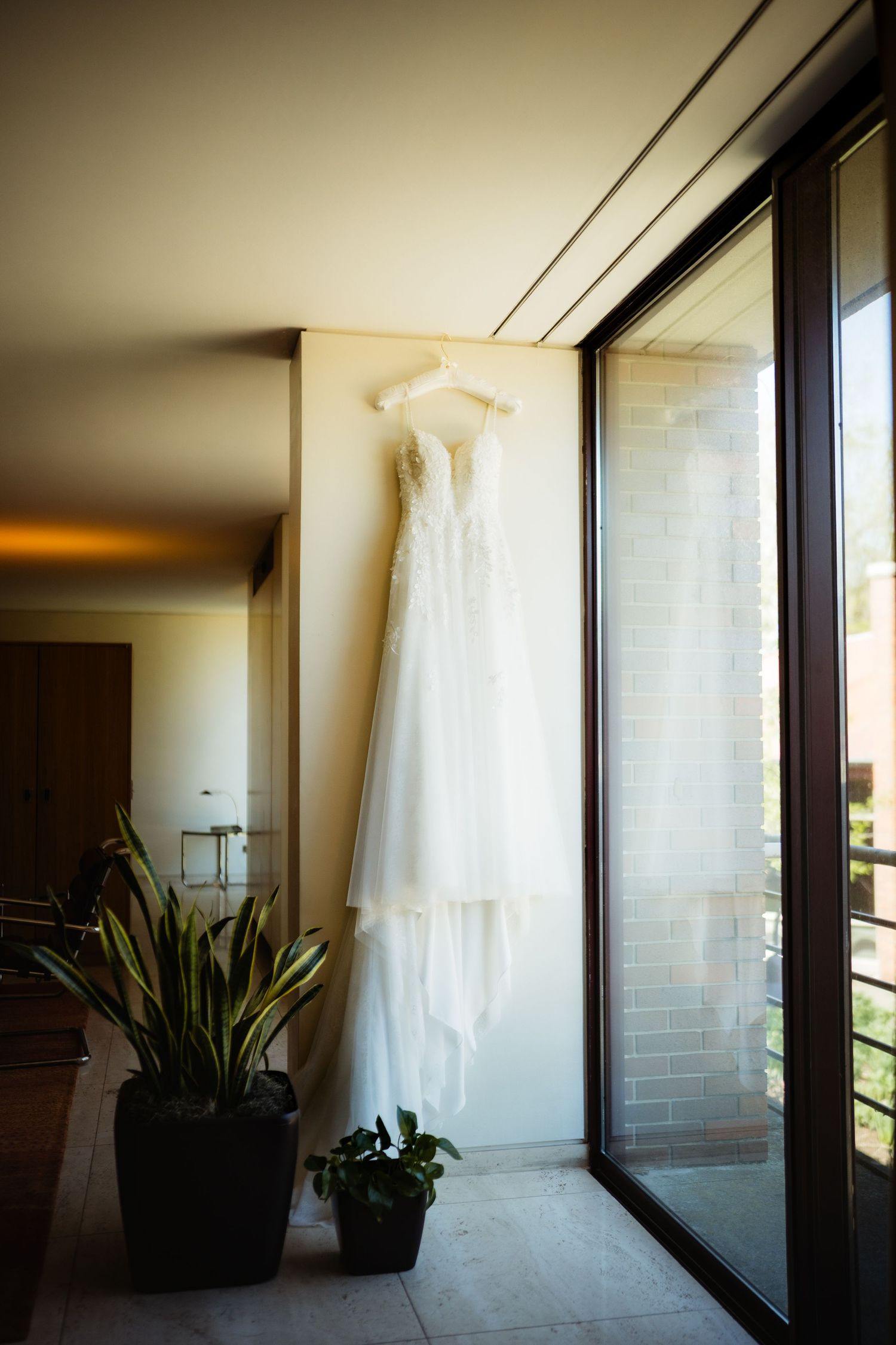 White wedding dress hanging on a hanger near a sunlit window with potted plants below.