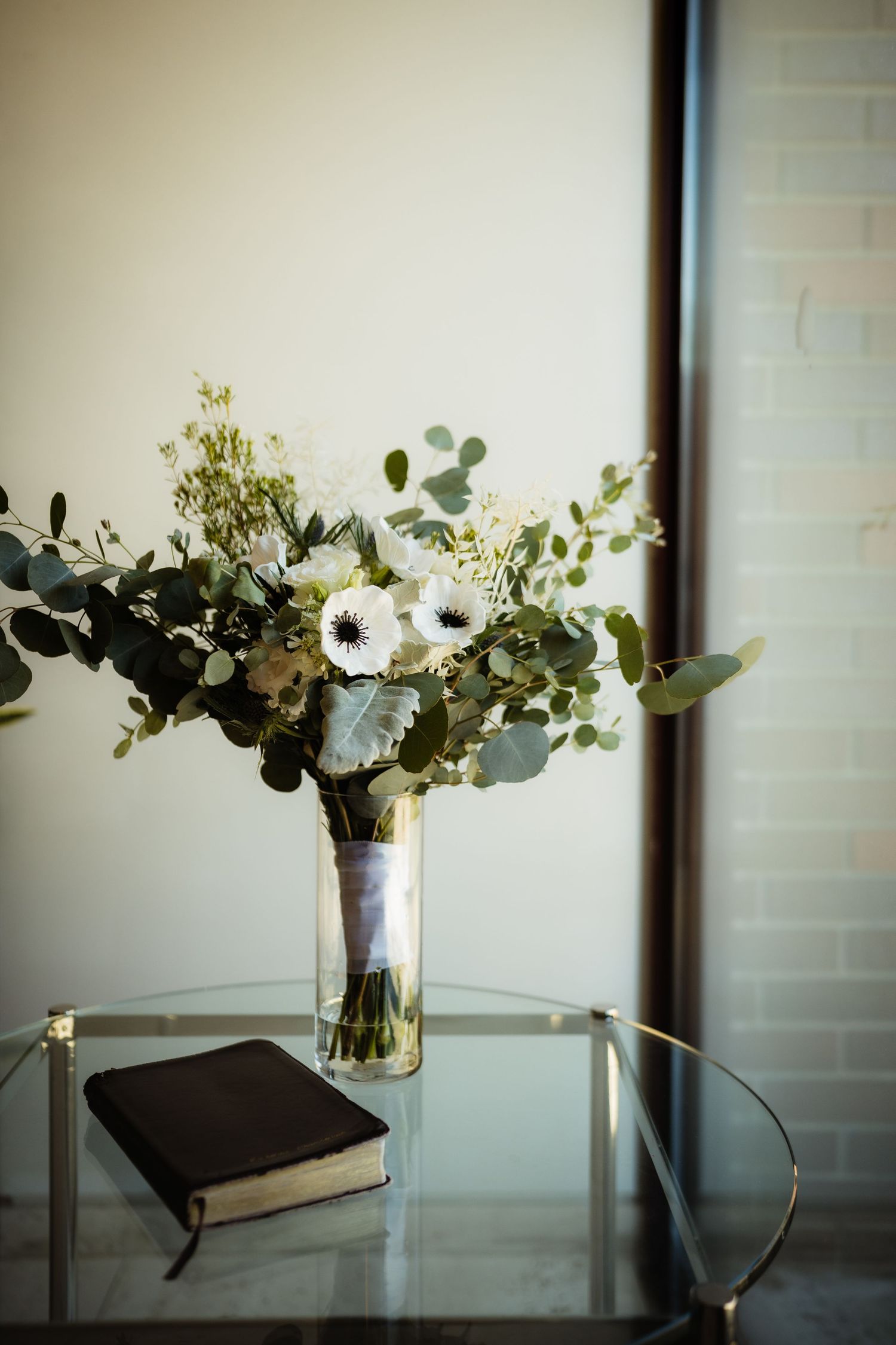 Elegant wedding bouquet with white anemones and eucalyptus in glass vase beside a black book on glass table.