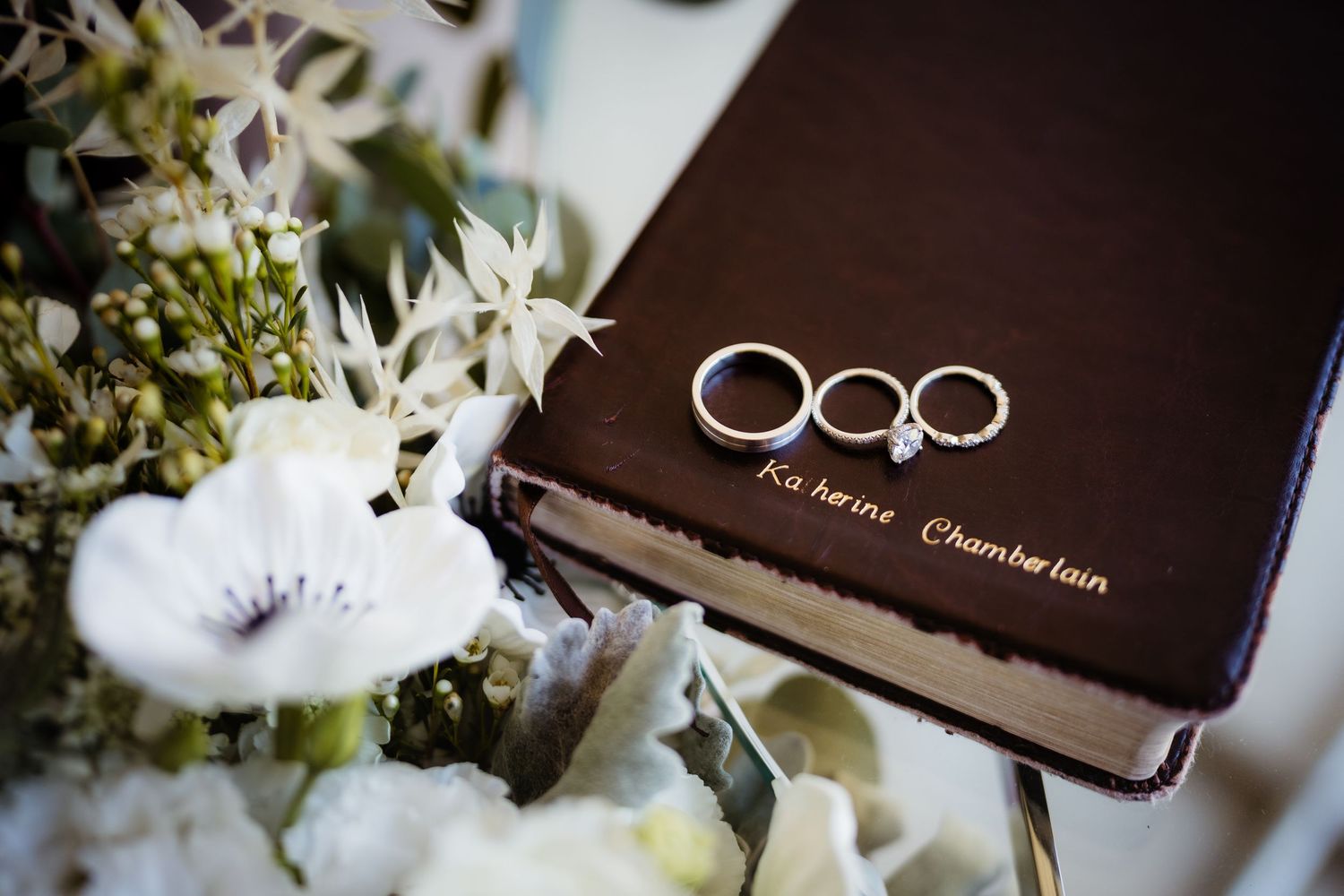 Wedding rings and engagement ring resting on a personalized dark leather Bible beside a white floral bouquet.